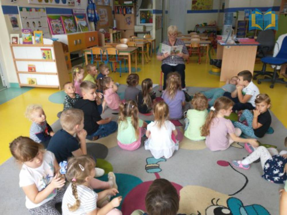 Zdjęcie: Dzieci w przedszkolu siedzą na dywanie, słuchają czytanej przez bibliotekarkę książki.